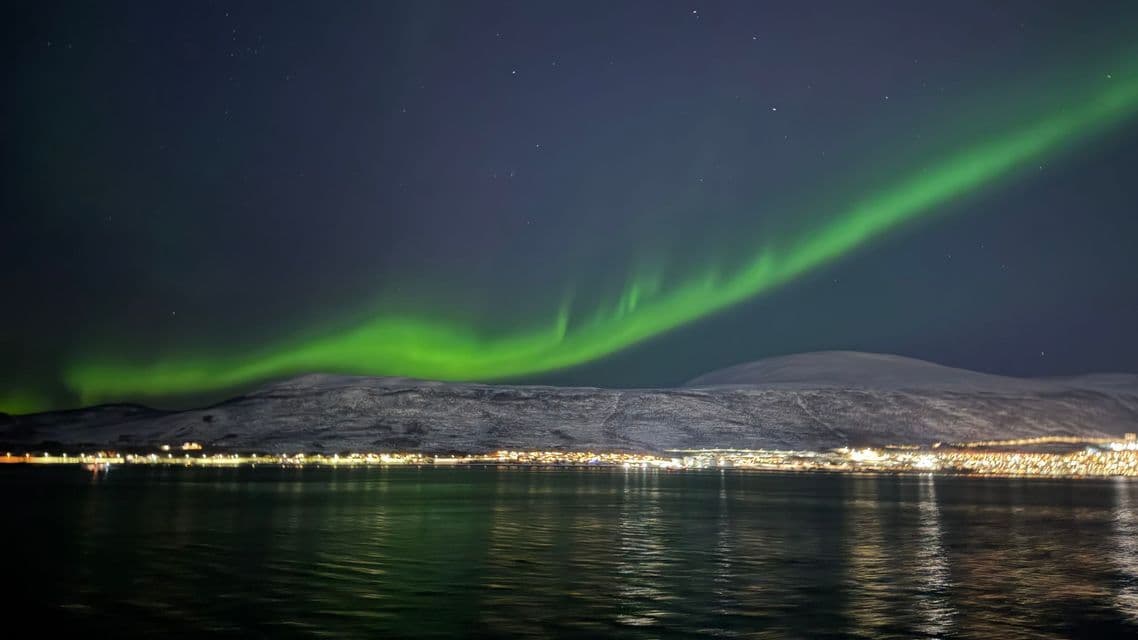 A band of green aurora borealis glows in the night sky over a snowy mountain and an illuminated coastal town reflected in the water.