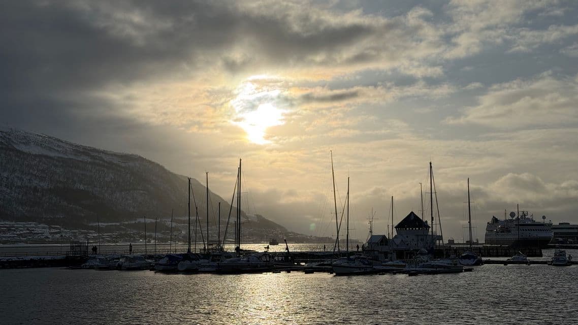 Sailboats are docked in a harbor at sunset, with light reflecting on the water and a snowy mountain in the background.