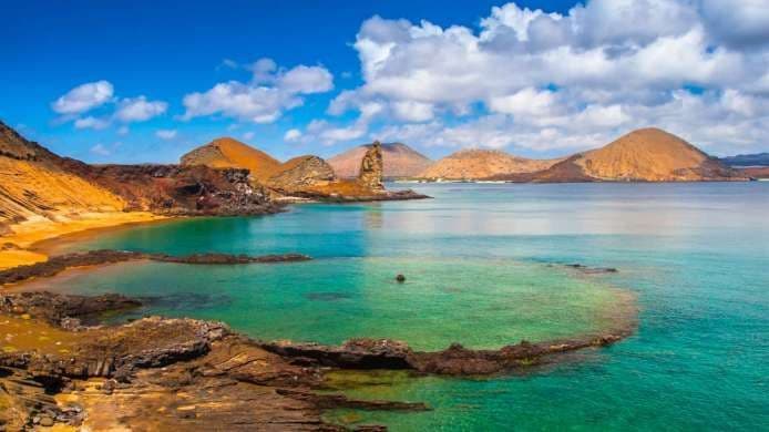 A volcanic coastline with a sandy beach meeting clear turquoise water, with distant hills under a partly cloudy blue sky.