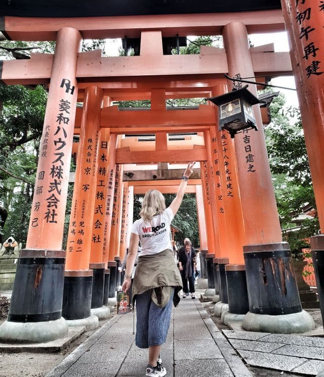 Un capogruppo WeRoad, visto di spalle, cammina lungo un sentiero di pietra attraverso un corridoio di torii arancioni.