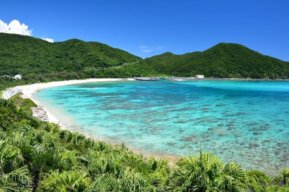 Una vista panoramica di una baia tropicale con una spiaggia di sabbia bianca, acqua cristallina turchese e rigogliose colline verdi sotto un cielo azzurro.