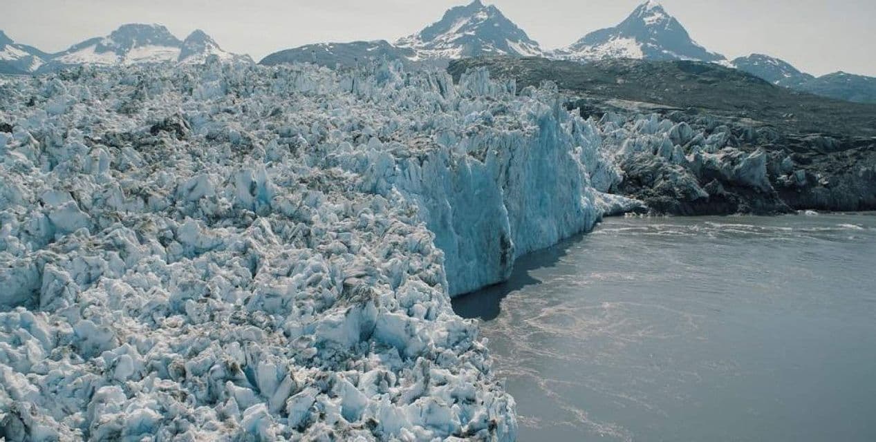 Un vasto ghiacciaio con una superficie fortemente crepacciata incontra uno specchio d'acqua di fronte a montagne innevate.