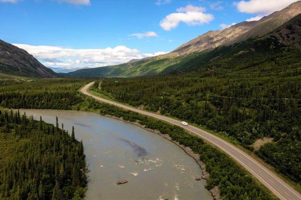 Una vista aerea di un camper bianco che guida su una strada tortuosa lungo un fiume attraverso una vasta valle montuosa e boscosa.