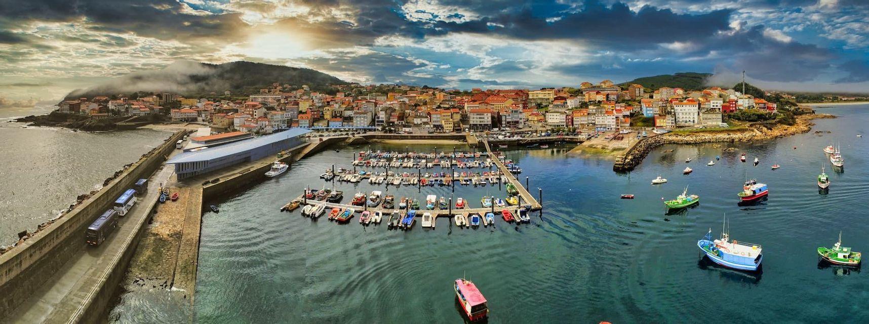 Una vista panorámica aérea de un colorido pueblo costero con un puerto lleno de barcos amarrados bajo un cielo nublado dramático.