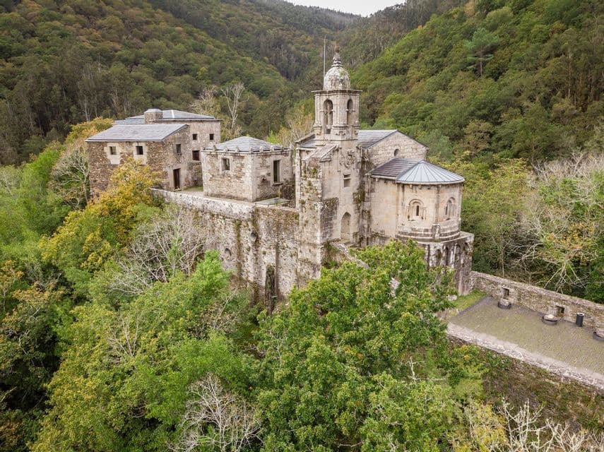 Una vista aérea de un antiguo monasterio de piedra con un campanario, enclavado en una ladera cubierta de densos árboles verdes.