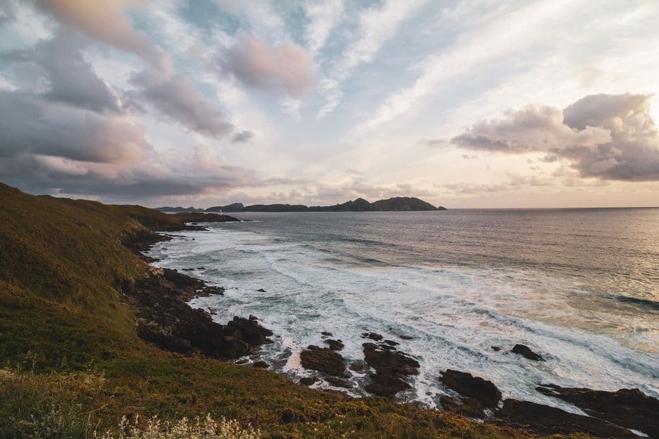 Una vista desde una colina cubierta de hierba de olas rompiendo contra una costa rocosa, con islas distantes en el horizonte bajo un cielo nublado al atardecer.