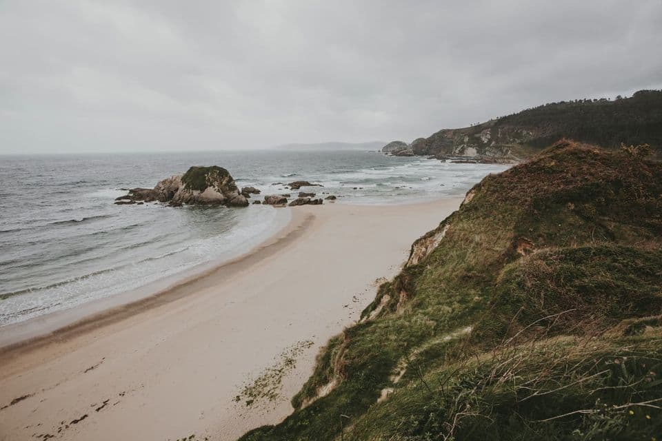Una vista desde un acantilado cubierto de hierba con vistas a una playa de arena con olas y afloramientos rocosos bajo un cielo nublado.