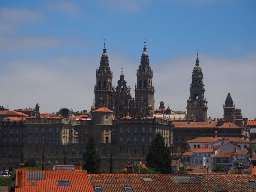 Una ornamentada catedral de piedra con múltiples agujas se eleva sobre los tejados de tejas rojas de una ciudad contra un cielo azul.