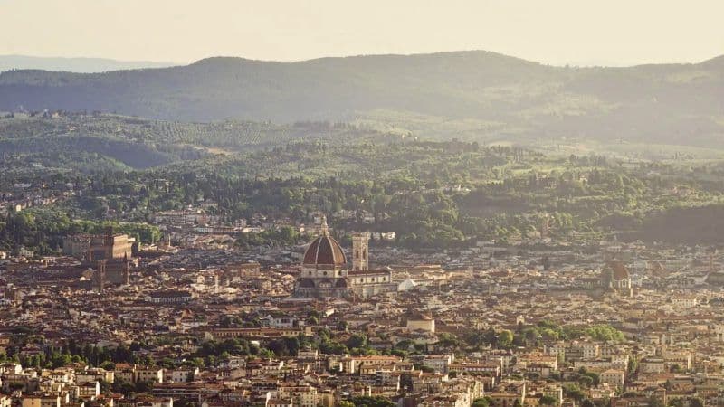 Vista panoramica di una storica città europea con una grande cattedrale a cupola, con dolci colline verdi sullo sfondo.