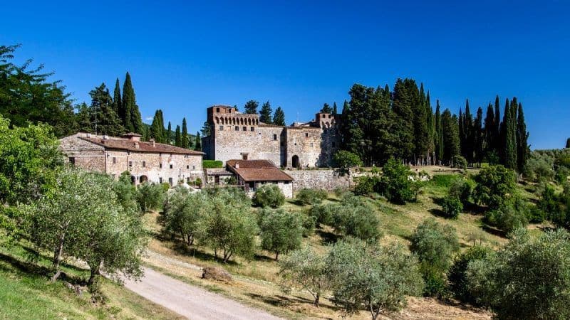 Un castello di pietra sorge su una verde collina, circondato da cipressi e ulivi, sotto un cielo azzurro e limpido.
