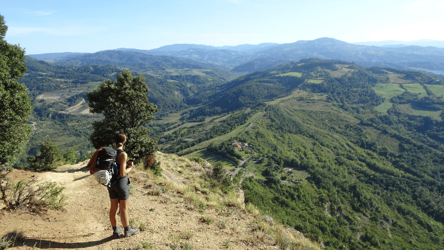 Un'escursionista con lo zaino si trova su un sentiero in cima a una scogliera, ammirando una vasta valle verde con colline ondulate.