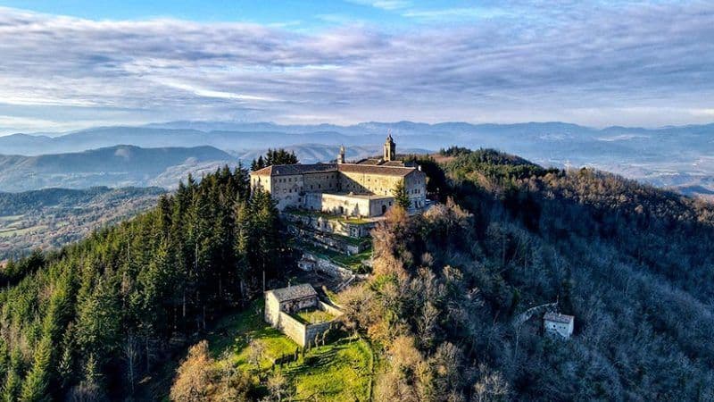 Una vista aerea di un grande complesso monastico in pietra arroccato su una cima montuosa boscosa, con colline ondulate sullo sfondo.