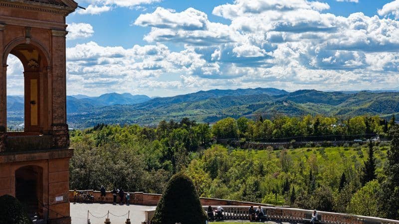 Vista dalla terrazza di un edificio che domina una valle di verdi colline ondulate e montagne sotto un cielo parzialmente nuvoloso.