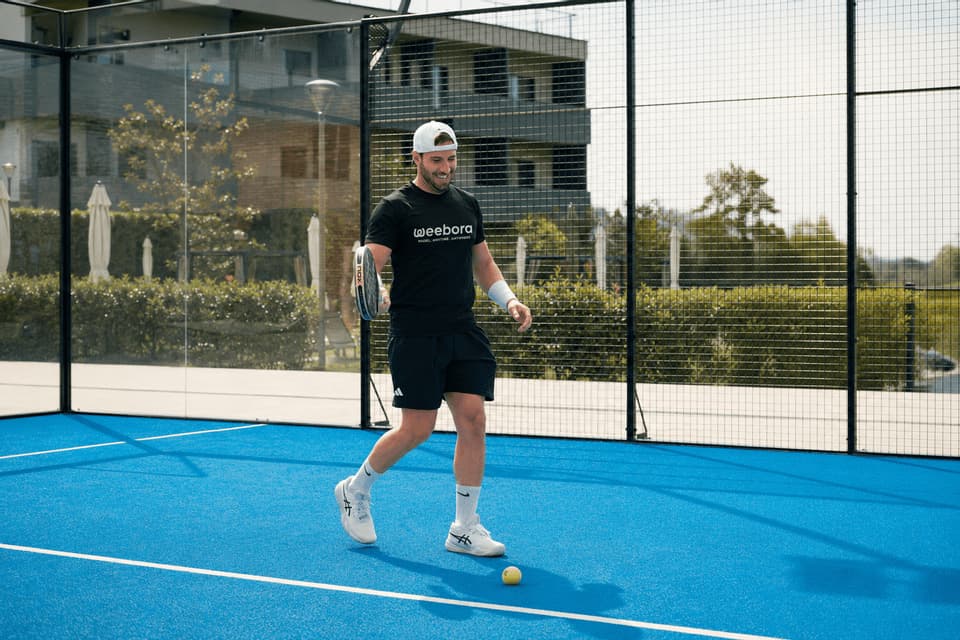 Un uomo sorridente, con maglietta nera e cappellino bianco, cammina su un campo da padel blu acceso, tenendo una racchetta.