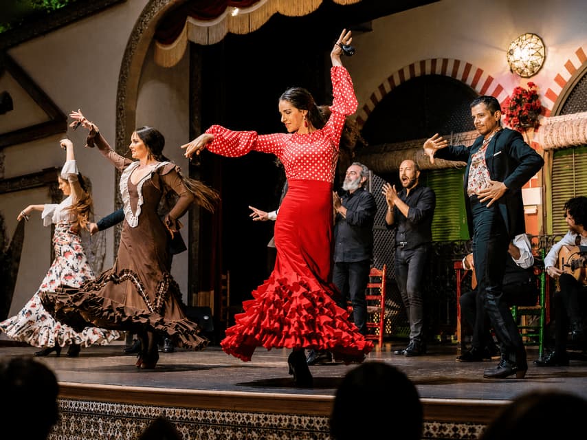 Una donna in un vestito a pois rossi si esibisce in flamenco su un palco, accompagnata da altri ballerini e musicisti.