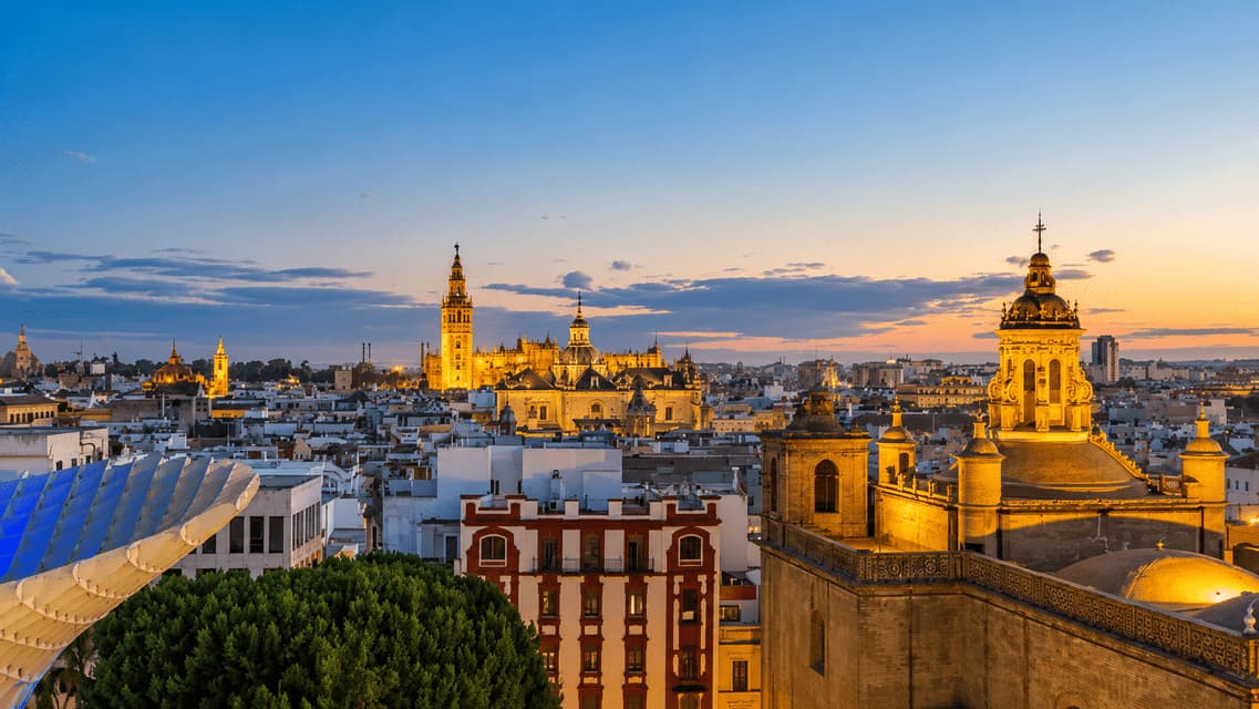 Una cattedrale e un campanile illuminati si ergono sopra i tetti di una città storica al tramonto.