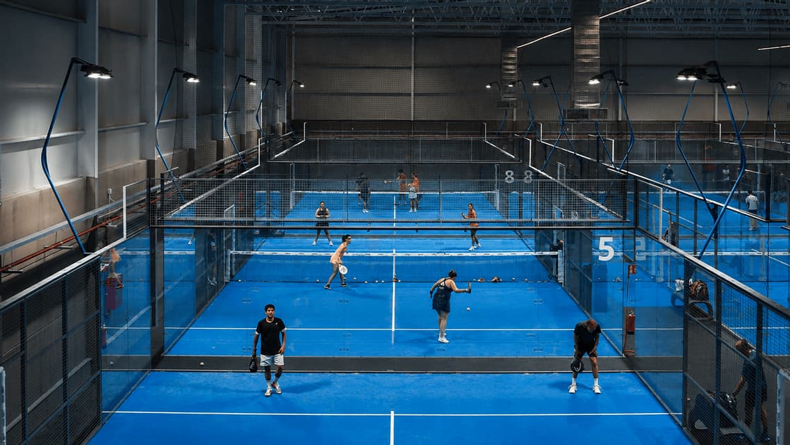 Una veduta dall'alto di persone che giocano a padel su diversi campi indoor con superfici blu brillanti, illuminati da grandi luci a soffitto.