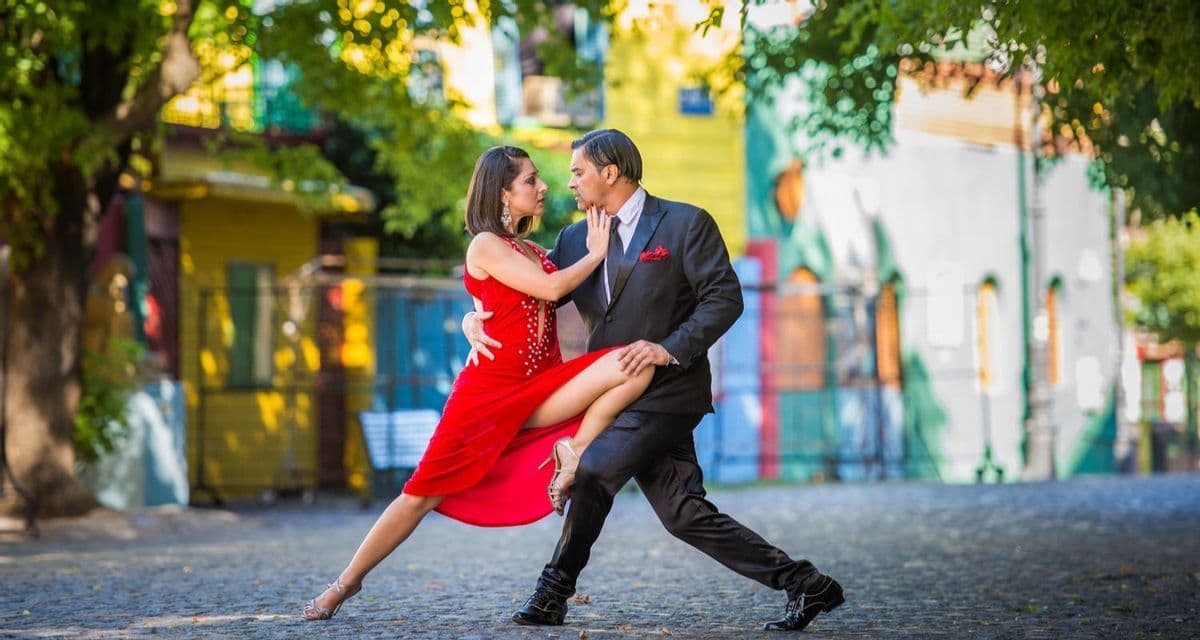 Un uomo e una donna in posa di tango su una strada acciottolata, con edifici colorati sullo sfondo.