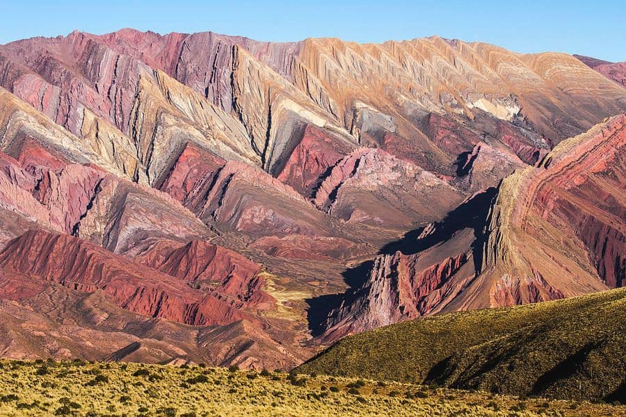 Una cadena montañosa muestra coloridas formaciones rocosas en capas en tonos de rojo, naranja y morado bajo un cielo azul claro.