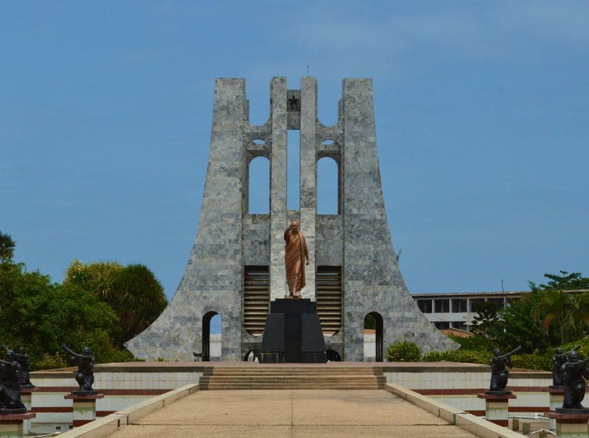 Una statua di bronzo si erge su un piedistallo davanti a un grande mausoleo in pietra grigia con un ampio viale che conduce ai suoi gradini sotto un cielo sereno.