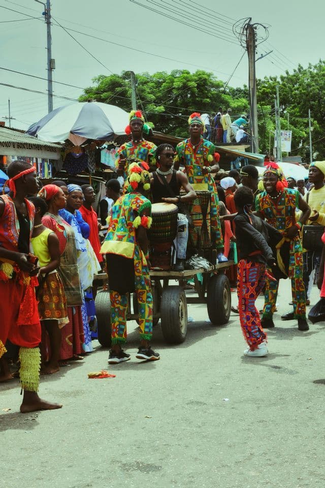 Un gruppo di musicisti in costumi vivaci suona i tamburi su un carro durante una parata di strada, circondato dalla folla.
