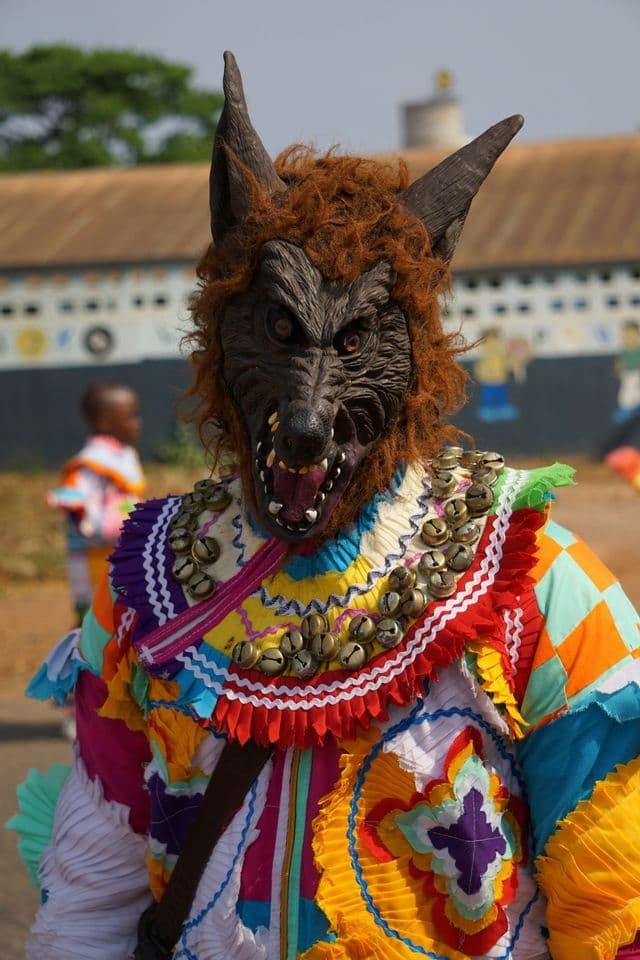 Una persona indossa un costume colorato e a più strati, caratterizzato da una minacciosa maschera da lupo con denti scoperti e un collare di campanelle.