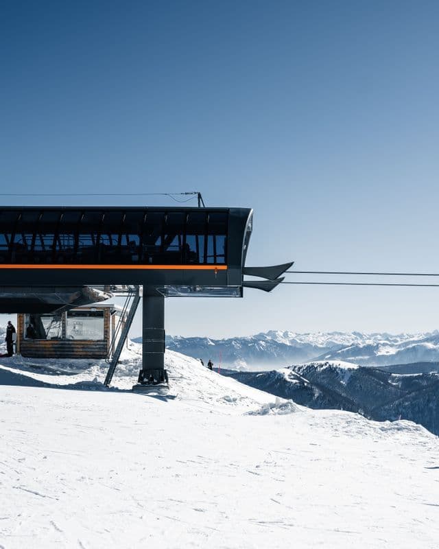 Una moderna stazione di risalita su una montagna innevata con cime distanti visibili sotto un cielo azzurro limpido.