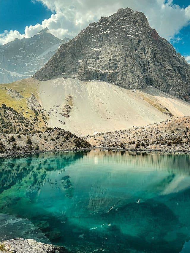 Un limpido lago alpino turchese riflette una grande montagna rocciosa sotto un cielo azzurro parzialmente nuvoloso.
