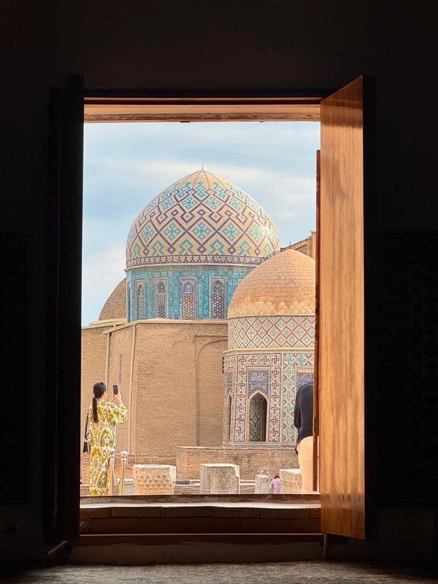 Inquadrata da un portale aperto, una donna fotografa un edificio con cupole ornate e piastrelle blu.