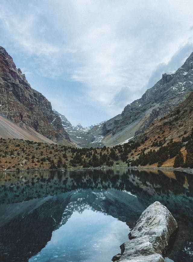 Un tranquillo lago alpino riflette le aspre montagne innevate di una valle sotto un cielo nuvoloso.