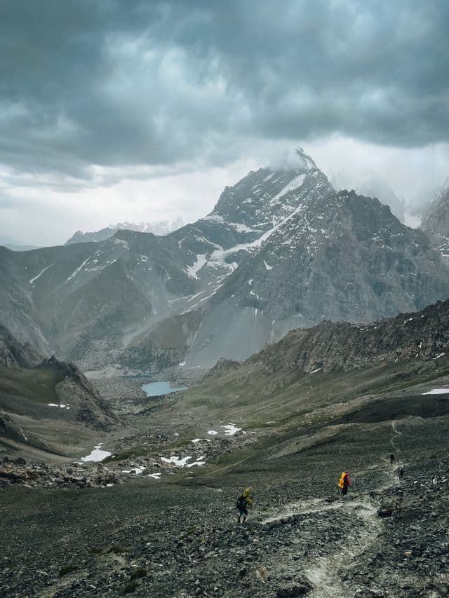 Un viaggio di gruppo WeRoad fa trekking su un sentiero roccioso attraverso una valle di montagna, con cime innevate e un lago turchese in lontananza.
