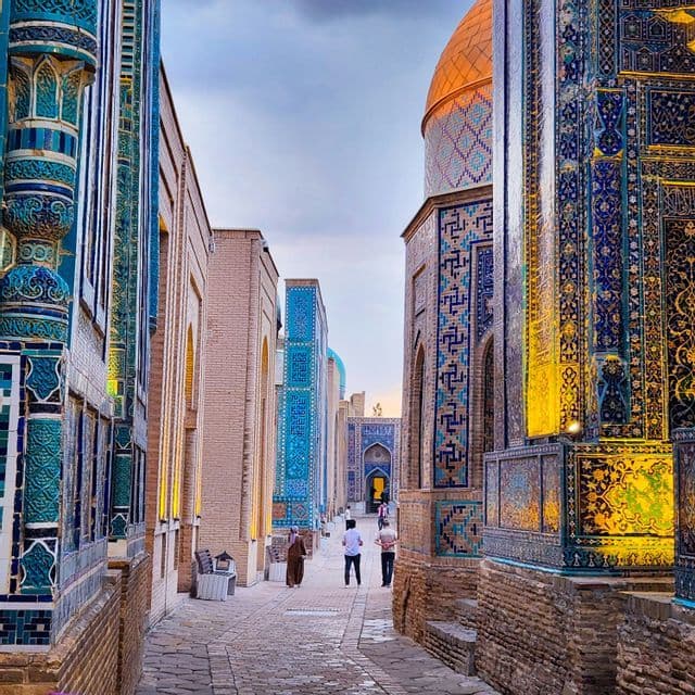 Un vicolo stretto e acciottolato fiancheggiato da edifici ornati con intricate piastrelle a mosaico blu, turchese e oro sotto un cielo nuvoloso.