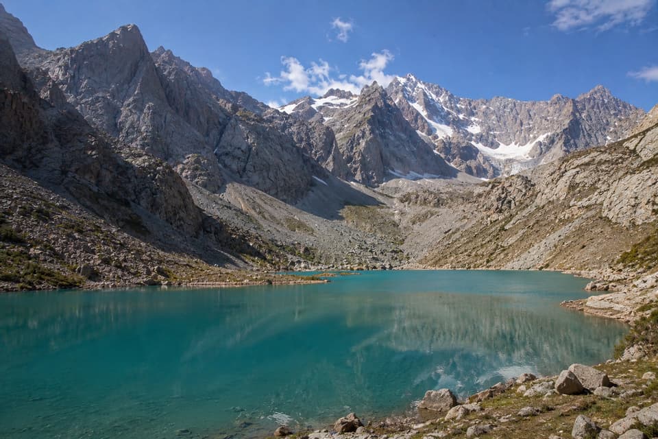 Un lago alpino turchese riflette alte montagne rocciose e innevate sotto un cielo azzurro.