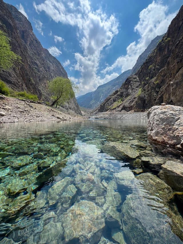 Una vista dal basso di un fiume cristallino in un canyon montano, con rocce sommerse visibili sotto un cielo azzurro e nuvole bianche.