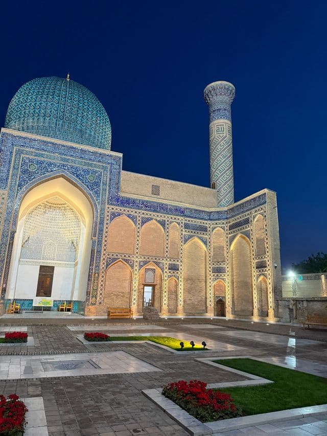 Una sontuosa moschea con cupola e minareto piastrellati di blu è illuminata da luci calde in un cortile di notte.
