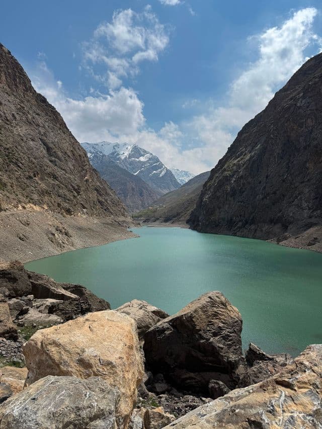 Un lago turchese si snoda attraverso un canyon ripido e roccioso, con montagne innevate in lontananza sotto un cielo parzialmente nuvoloso.