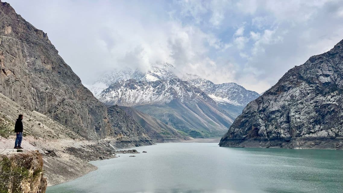 Un uomo si trova sul bordo di una scogliera che si affaccia su un lago circondato da montagne rocciose e innevate, sotto un cielo nuvoloso.