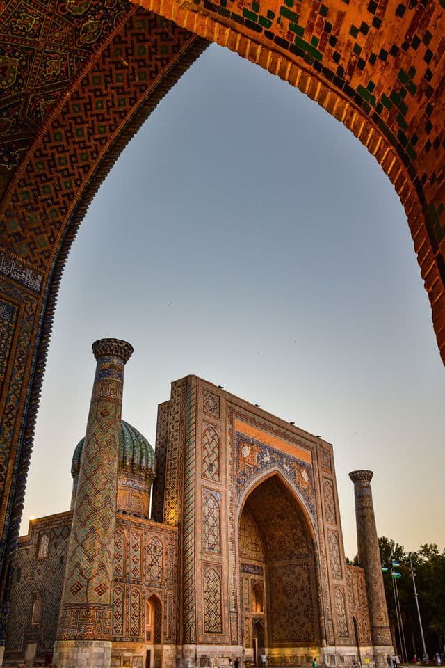 Veduta dal basso di un edificio ornato e piastrellato con minareti, incorniciato da un grande arco in mattoni contro un cielo limpido al tramonto.
