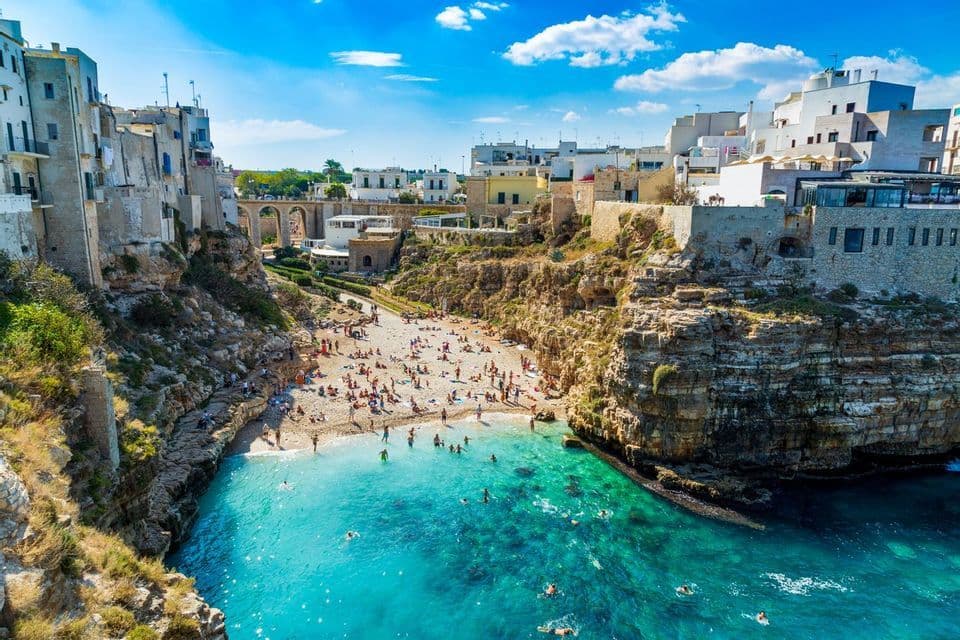 Una veduta aerea di una spiaggia affollata in una cala rocciosa, con persone che nuotano nell'acqua turchese ed edifici arroccati sulle scogliere circostanti.