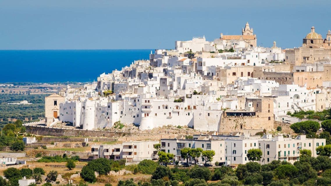 Una città bianca adagiata su una verde collina, affacciata sul mare blu cobalto sotto un cielo limpido.