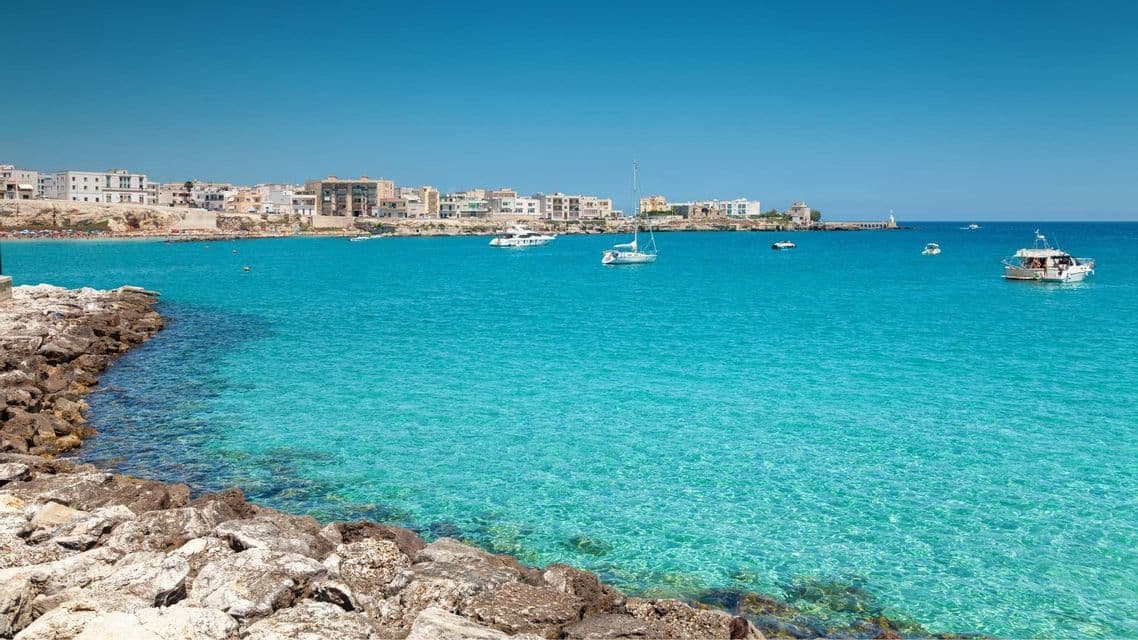 Vista da una costa rocciosa di una località marittima, con una baia dalle acque limpide e turchesi e diverse imbarcazioni.