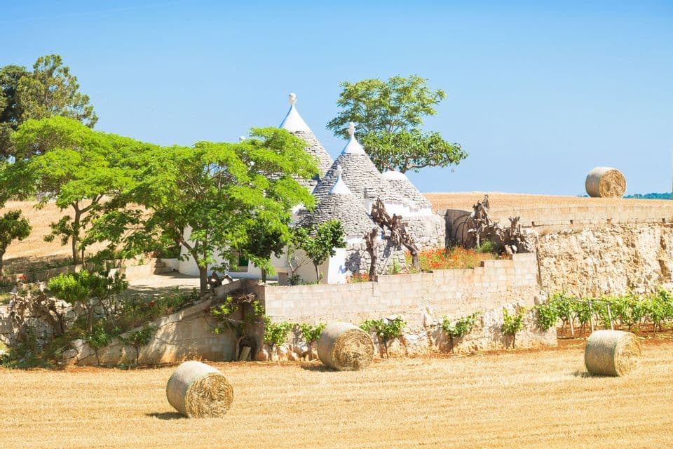 Trulli bianchi con tetti conici in pietra si ergono su una collina verde sopra un campo mietuto con balle di fieno rotonde.