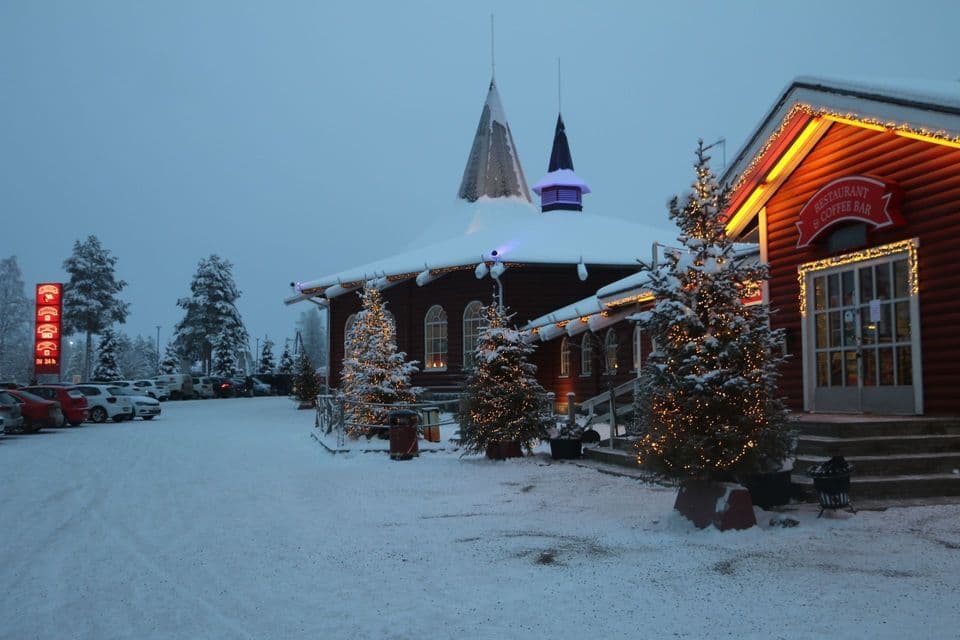 Un ristorante e caffetteria in legno, con tetti innevati e luci natalizie, si trova in un paesaggio innevato accanto ad alberi decorati.