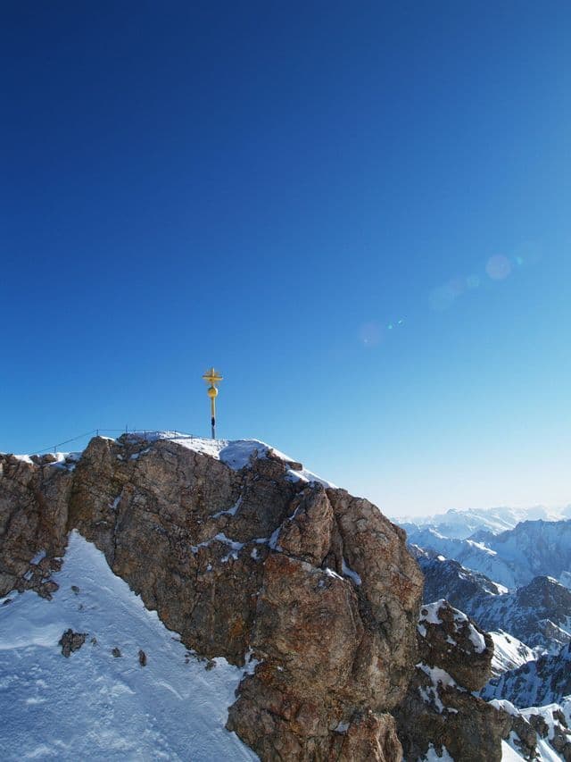 Ein gelbes Gipfelkreuz steht auf einem felsigen, schneebedeckten Berggipfel unter einem weiten, klaren blauen Himmel mit Lens Flare.