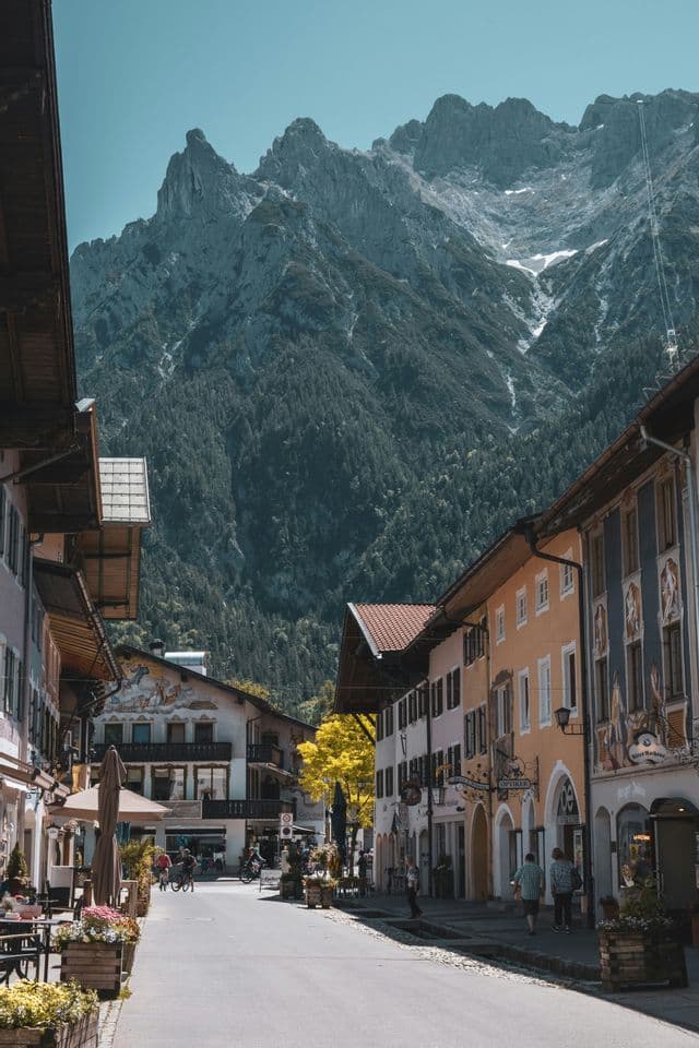 Eine von traditionellen Gebäuden gesäumte Straße in einer Bergstadt, mit einem aufragenden, felsigen Gipfel im Hintergrund unter klarem Himmel.