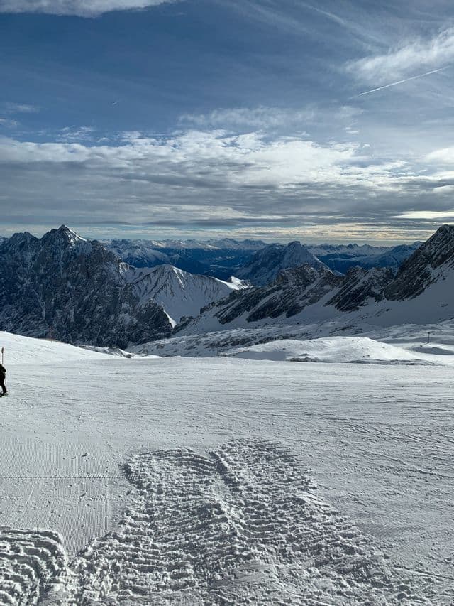 Ein Panoramablick von einer präparierten Skipiste über eine weite Kette schneebedeckter Berge unter bewölktem Himmel.