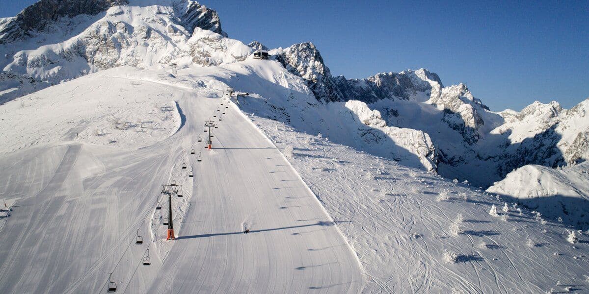 Ein einsamer Skifahrer fährt auf einer breiten, präparierten Skipiste einen weitläufigen, schneebedeckten Berg hinab, unter klarem Himmel und neben einem Sessellift.
