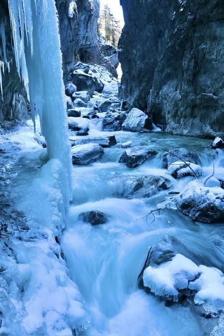 Ein schnell fließender Fluss rauscht durch eine enge, eisige Schlucht, gesäumt von schneebedeckten Felsen und herabhängenden Eiszapfen.