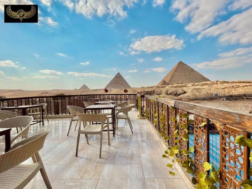 An outdoor terrace with empty tables and chairs overlooking several large pyramids in the desert under a partly cloudy sky.