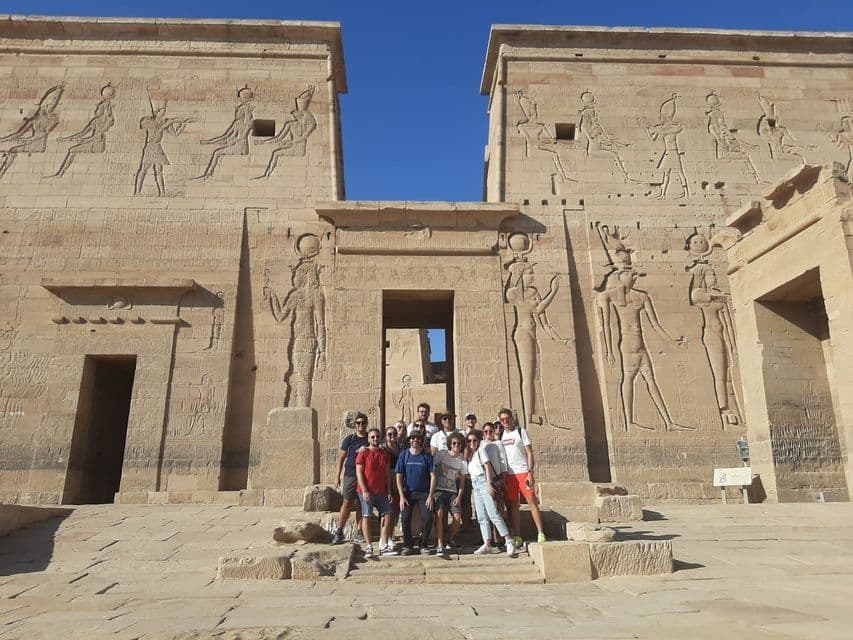 A WeRoad group trip poses on the stone steps of an ancient Egyptian temple with large carved hieroglyphs.